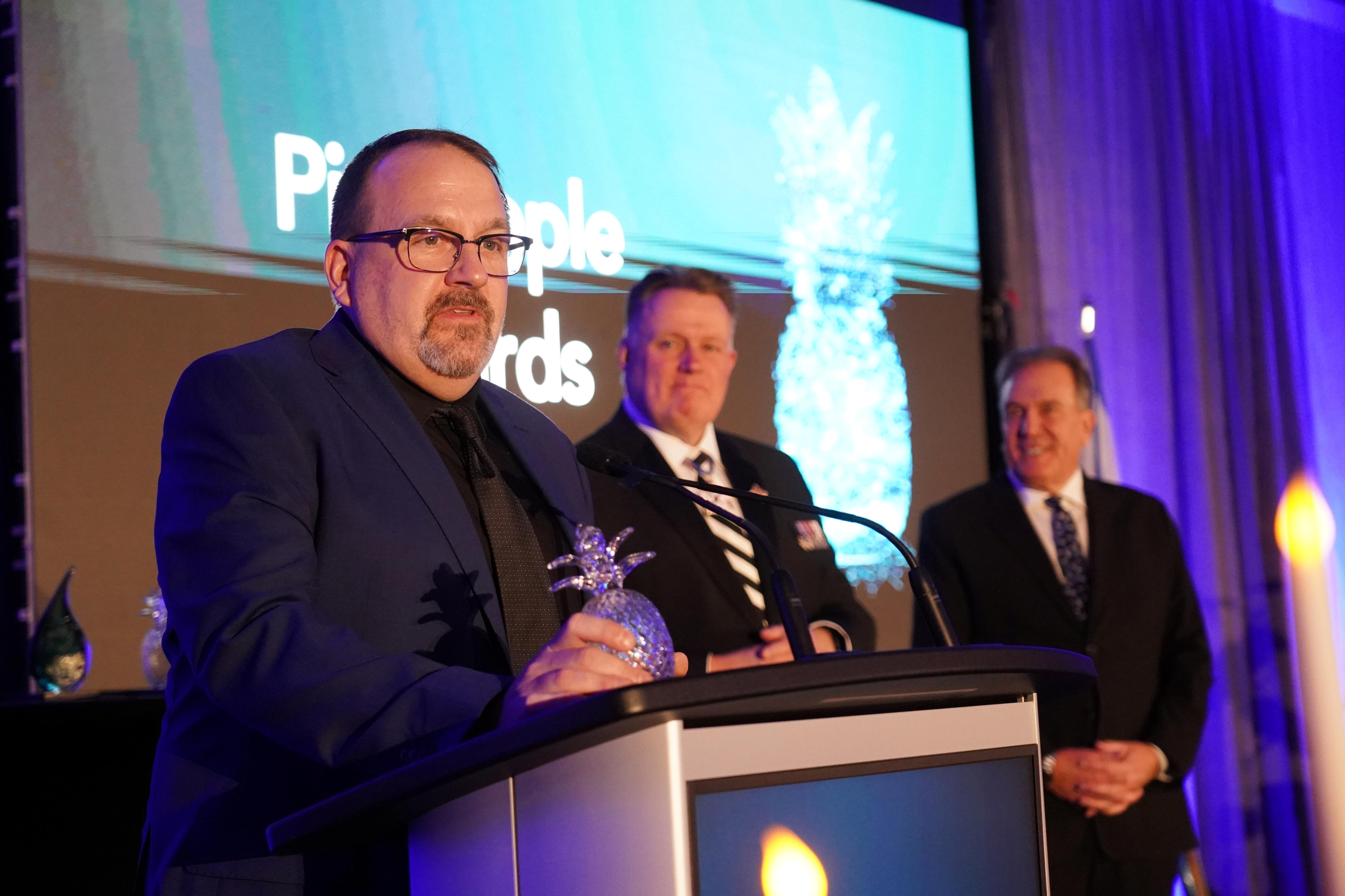 A man stands at a podium accepting an award while two other men stand in the background to the right.